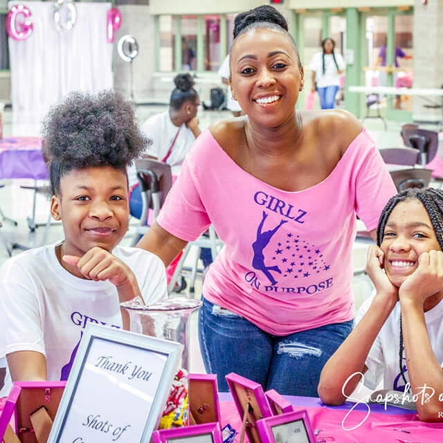A woman stands behind sitting two girls; they are all smiling at the Girlz on Purpose Event